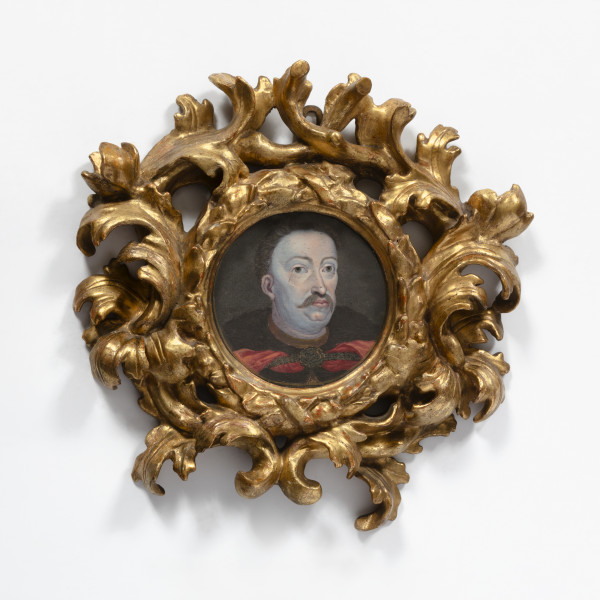 Fotografia kolorowa. Owalna rama w kolorze złotym. W otoku ozdobiona motywem liści, które rozchodzą się tworząc kolejny krąg z tym samym motywem. Pośrodku portret mężczyzny w średnim wieku. Twarz zdecydowanie okrągła, blada. Duże oczy i wyraźnie zarysowane łuki brwiowe. Usta wydatne, a nad nimi okazałe wąsy. Włosy ciemne, zaczesane do góry, odsłaniają wysokie czoło. Ramiona mężczyzny okryte czarnym kołnierzem, spod którego widać fragment ubrania w kolorze czerwonym. Pośrodku, na piersi okrągła zapinka z rozchodzącymi się na boki paskami.