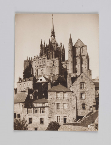 MC/M/38/34/ML - Fotografia w pozycji wertyalnej. Linia horyzontu dzieli zdjęcie na mniej więcej połowę. Centrum obrazu przesunięte nieco na lewą stronę. Opactwo wraz z przylegającymi budynkami kompozycyjnie wpisane w trójkąt. Światło rozproszone, jego źródło znajduje się poza fotografią z lewej strony.
Na zdjęciu ukazano zabudowania Mont Saint-Michel od strony wschodniej. Na pierwszym planie, poniżej linii horyzontu znajdują się zabudowania wioski. Powyżej linii horyzontu znajduje się opactwo wraz z blankami - po prawej stronie tak zawany La Merveille z refektarzem, po lewej zaś absyda katedry z iglicą zbudowaną na przecięciu naw.
Na odwrocie (ustawienie w pozycji horyzontalnej) w prawym górnym rogu napisana niebieskim długopisem liczba 