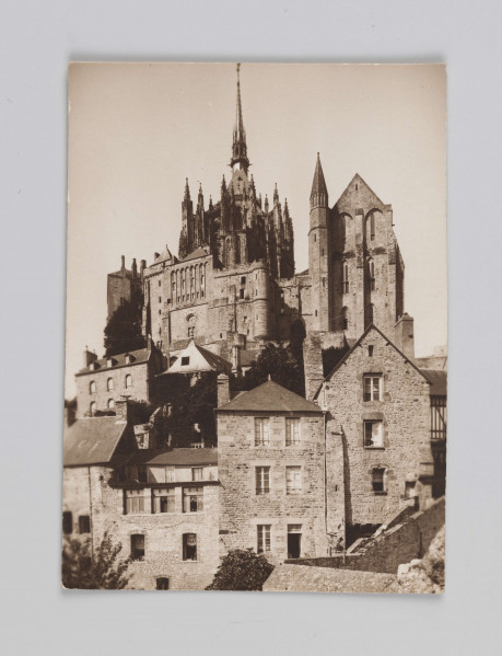 MC/M/38/34/ML - Fotografia w pozycji wertyalnej. Linia horyzontu dzieli zdjęcie na mniej więcej połowę. Centrum obrazu przesunięte nieco na lewą stronę. Opactwo wraz z przylegającymi budynkami kompozycyjnie wpisane w trójkąt. Światło rozproszone, jego źródło znajduje się poza fotografią z lewej strony.
Na zdjęciu ukazano zabudowania Mont Saint-Michel od strony wschodniej. Na pierwszym planie, poniżej linii horyzontu znajdują się zabudowania wioski. Powyżej linii horyzontu znajduje się opactwo wraz z blankami - po prawej stronie tak zawany La Merveille z refektarzem, po lewej zaś absyda katedry z iglicą zbudowaną na przecięciu naw.
Na odwrocie (ustawienie w pozycji horyzontalnej) w prawym górnym rogu napisana niebieskim długopisem liczba 