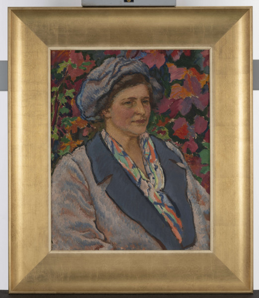 Portret ujęty do pasa. Postać zwrócona w 3/4. Kobieta ubrana w niebieski beret, kolorową bluskę widoczną spod niebieskiego żakietu z szafirowym kołnierzem. Twarz okrągła. Oczy wąskie, nos długi, usta wyraźnie zaznaczone. Tło stanowią czerwone, pomarańczowe i fioletowe kwiaty z zielonymi liśćmi.
