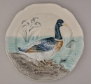 Talerz fajansowy, płytki o łagodnie podniesionym brzegu, wykrojonym w sześć płaskich łuków. Dekoracja reliefowa wklęsło-wypukła przedstawia kaczkę stojącą na niewielkim cyplu wśród wodnego krajobrazu