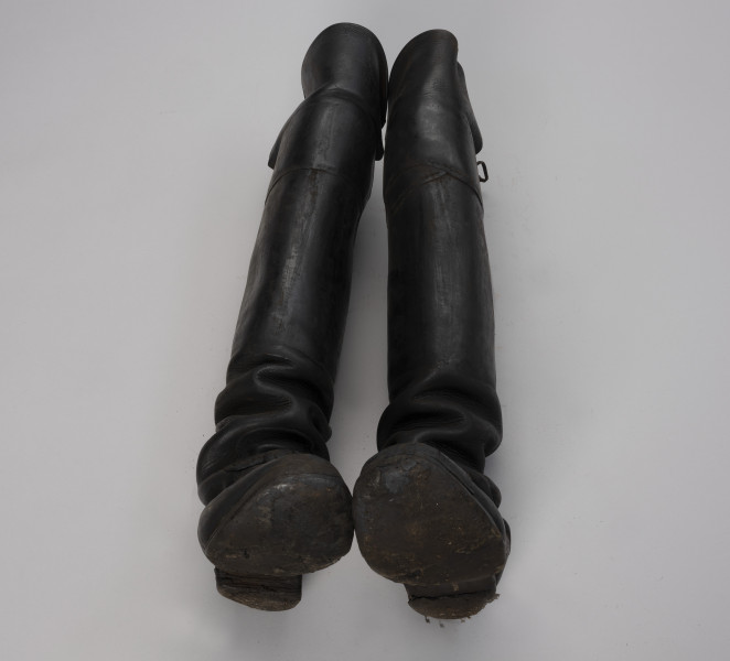 tłok pieczętny - Ujęcie z przodu; Para butów rybackich o wysokich cholewach zszytych z dwóch kawałków skóry, które od przodu sięgają powyżej kolan (ochraniacze). Drewniane podeszwy są podbite grubą, barwioną na kolor czarny, sztywną, prawdopodobnie impregnowaną tłuszczem, skórą za pomocą gwoździków. Czubek prawego buta jest wzmocniony kawałeczkiem skóry, a lewy ma wzmocnione skórą palce.