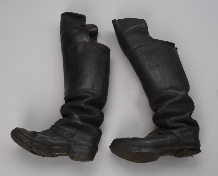 tłok pieczętny - Ujęcie z drugiego boku; Para butów rybackich o wysokich cholewach zszytych z dwóch kawałków skóry, które od przodu sięgają powyżej kolan (ochraniacze). Drewniane podeszwy są podbite grubą, barwioną na kolor czarny, sztywną, prawdopodobnie impregnowaną tłuszczem, skórą za pomocą gwoździków. Czubek prawego buta jest wzmocniony kawałeczkiem skóry, a lewy ma wzmocnione skórą palce.