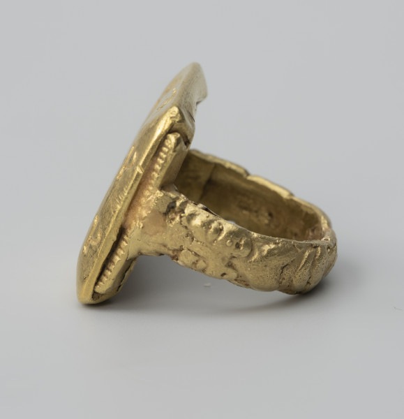 pierścień z Pęzina - Ujęcie z boku; Pierścień zbudowany z obrączki i dużej prostokątnej tarczki. Zewnętrzna część obrączki, lekko wypukła, ukształtowana została w formie dwóch antytetycznie ułożonych lwów dotykających paszczami tarczki. Tarczka prostokątna, zbliżona do kwadratu, o fasetowanych bokach. Wzdłuż boków łączących się z obrączką, wąskie strzemiączka ozdobione nacinaniem.