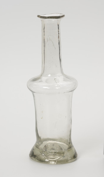 karafinka - Ujęcie z przodu; Niewielka butelka o pojemności 0,25 L. Wydmuchana na piszczelu z bezbarwnego szkła – na brzuścu widoczne smugi z nie do końca prawidłowego procesu produkcji. Kształt podłużny, wąski, o grubym, wyraźnie wyodrębnionym dnie, brzusiec w górnej części uwypuklony przechodzi w wąską, długą szyjkę zakończoną płasko wywiniętym niewielkim wylewem. Na obwodzie podstawy w formie odciśniętego, owalnego stempla umieszczony napis: 