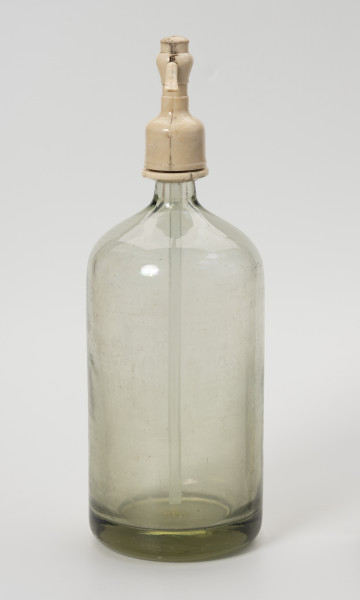 syfon do wody sodowej - Ujęcie z lewej; Grubościenna szklana butla o pogrubionym dnie i krótkiej, wąskiej szyjce z bezbarwnego szkła zwieńczona zamknięciem wykonanym z jasnego tworzywa sztucznego, składającego się z kraniku niewidocznego na ujęciu i ruchomej rączki połączonej ze szklaną rurką wpuszczoną do wnętrza butli. Po naciśnięciu rączki zamknięcia, leci woda gazowana.