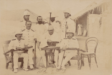 fotografia z albumu Aleksandra Dandelskiego; Aleksander Dandelski w towarzystwie członków ekspedycji wojskowej do Niemieckiej Afryki Wschodniej - ujęcie z przodu; Fotografia w układzie poziomym przedstawia ośmiu mężczyzn ustawionych w dwóch rzędach. W pierwszym czterech z nich siedzi na wyplatanych w stylu kolonialnym fotelach oraz krześle (pierwszy po prawej to Aleksander Dandelski), w drugim kolejnych czterech stoi. Wszyscy ubrani są na biało. Większość ma na sobie mundur, prawdopodobnie oficerski lub elegancki strój. Część z nich ma na głowach białe kolonialne hełmy, a w dłoniach trzyma laski. Przed mężczyznami stoi kawiarniany stolik, na którym postawiono hełm. Po prawej stronie pozostawiono puste krzesło do którego przypięto łańcuchem jeden z foteli. U stóp siedzących mężczyzn leży śpiący pies. W tle widać miejscową kobietę i tubylczą zabudowę.