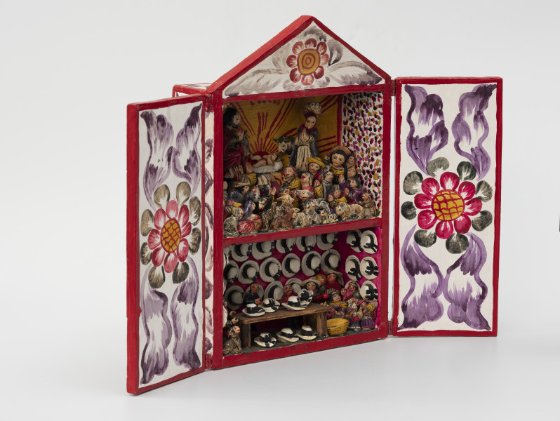 retablo - Ujęcie ze skosu z lewej; Jasna, bogato dekorowana kolorowymi wzorami florystycznymi, prostopadłościenna, wewnątrz dwupoziomowa skrzynka (kapliczka) z dwuskrzydłowymi drzwiczkami, zwieńczona trójkątną koroną. Na wyższym poziomie pokazano Narodzenie Dzieciątka Jezus, gdzie wykonane z pasty ziemniaczanej i gipsu, polichromowane postacie Marii, Józefa i malutkiego Jezusa ustawiono na podwyższeniu. Bezpośrednio za Świętą Rodziną znajdują się zwierzęta: osioł i krowa. Poniżej głównych (nieco większych od pozostałych) figur zaprezentowano miejscową ludność (kobiety, mężczyzn i dzieci) w pozycji modlitewnej – większość klęczy z dłoniami złożonymi i skierowanymi ku Dzieciątku. Pierwszy rząd wypełniają najmniejsze figurki zwierząt hodowlanych: kóz, owiec, cieląt, jagniąt, krów, kur, źrebiąt oraz kota. Tło przedstawienia składa się z dwóch różnych dekoracji. Po obu bokach zastosowano wzór kropkowy (wielokolorowy) na białym tle, natomiast centralnie za postaciami namalowano usytuowane nisko na horyzoncie czerwone słońce na żółtym tle. Dolne piętro przedstawia scenę rodzajową: sprzedawcę kapeluszy w sklepie lub na targu w towarzystwie miejscowej ludności, instrumentów muzycznych i prawdopodobnie psa. Różowe tło wypełniono trzema rzędami figurek przedstawiających biało-czarne kapelusze typu panama.