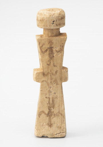 figurka magiczna - Ujęcie z tyłu; Stojąca figurka postaci ludzkiej o zredukowanej formie wykonana z jasnego drewna. Głowę nieznacznie oddzielono od tułowia wcięciem imitującym szyję. Na długim, wykonanym schematycznie korpusie w kształcie klepsydry zaznaczono jedynie ręce lub dłonie. Całość pokryto geometrycznymi (linie, łuki, kropki) i antropomorficznymi malunkami wykonanymi wyblakłym już czarnym barwnikiem. Rysunki wykonane na tułowiu ułożono na osi pionowej. Na jasnej powierzchni drewna widoczne są większe i mniejsze spękania, ślady ciosania, malutkie otworki i oznaczenia badacza w postaci: literki 