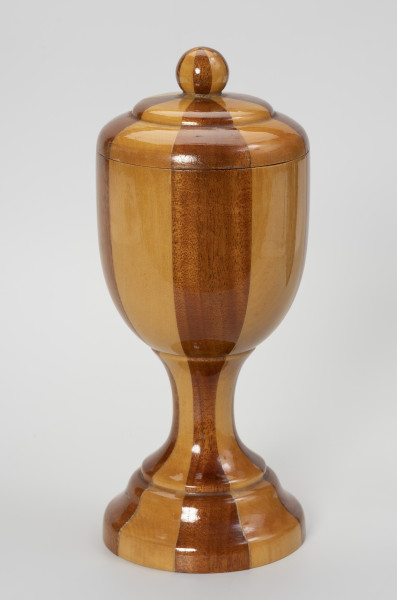puchar z pokrywką - ujęcie z przodu; Pamiątkowy drewniany puchar ze zdejmowaną pokrywką, zakończoną okrągłym uchwytem. Obrobiony na tokarce z drewnianego klocka klejonego z koncentrycznie ułożonych kawałków drewna w dwóch odcieniach – jasno-żółtawym i ciemno-brązowym – ułożonych naprzemiennie. Pokrywa uformowana na wzór wycinka spłaszczonej, podwójnej bańki z gałką na szczycie. Czara o formie niewysokiego cylindra o zaokrąglonym dnie osadzona jest na kolumnowej, przewężonej nóżce o stopie w formie ściętego stożka.