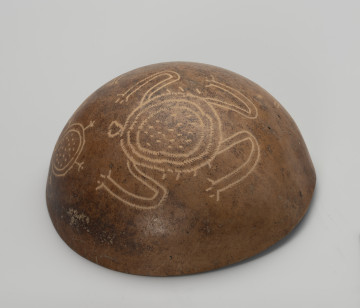 Naczynie do serwowania jedzenia, napojów i guarapo - Ujęcie z przodu, spód misy; Miska z tykwy o naturalnym jasnobrązowym, orzechowym kolorze z zewnętrzną powierzchnią bogato zdobioną rytymi ornamentami zoomorficznymi i jednym antropomorficznym. Ornamentacja wykonywana za pomocą metalowego narzędzia, jego ślady w postaci płytkich i drobnych nacięć widoczne są na wyrytych rysunkach przedstawiających trudne do zidentyfikowania zwierzęta. Największy z nich to najprawdopodobniej żółw lądowy w towarzystwie mniejszego żółwia. Na zewnętrznej powierzchni tykwy widoczne są mniejsze nacięcia i wgłębienia, przetarcia oraz ślady użytkowania.