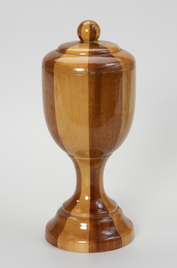 puchar z pokrywką - ujęcie z przodu; Pamiątkowy drewniany puchar ze zdejmowaną pokrywką, zakończoną okrągłym uchwytem. Obrobiony na tokarce z drewnianego klocka klejonego z koncentrycznie ułożonych kawałków drewna w dwóch odcieniach – jasno-żółtawym i ciemno-brązowym – ułożonych naprzemiennie. Pokrywa uformowana na wzór wycinka spłaszczonej, podwójnej bańki z gałką na szczycie. Czara o formie niewysokiego cylindra o zaokrąglonym dnie osadzona jest na kolumnowej, przewężonej nóżce o stopie w formie ściętego stożka.