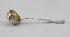 łyżka wazowa - Ujęcie z boku; Rokokowa łyżka wazowa o puklowanej powierzchni i falistym brzegu, z rytowaną podwójną linią przy krawędzi. Pukle wydzielone czterema parami repusowanych esowatych linii. Wnętrze czerpaka złocone. Trzonek płaski, o asymetrycznym, falistym wykroju. Uchwyt wydzielony przewężeniem, na końcu rozszerzający się i wygięty lekko w górę, zdobiony grawerowaną i fakturowaną rokokową dekoracją.