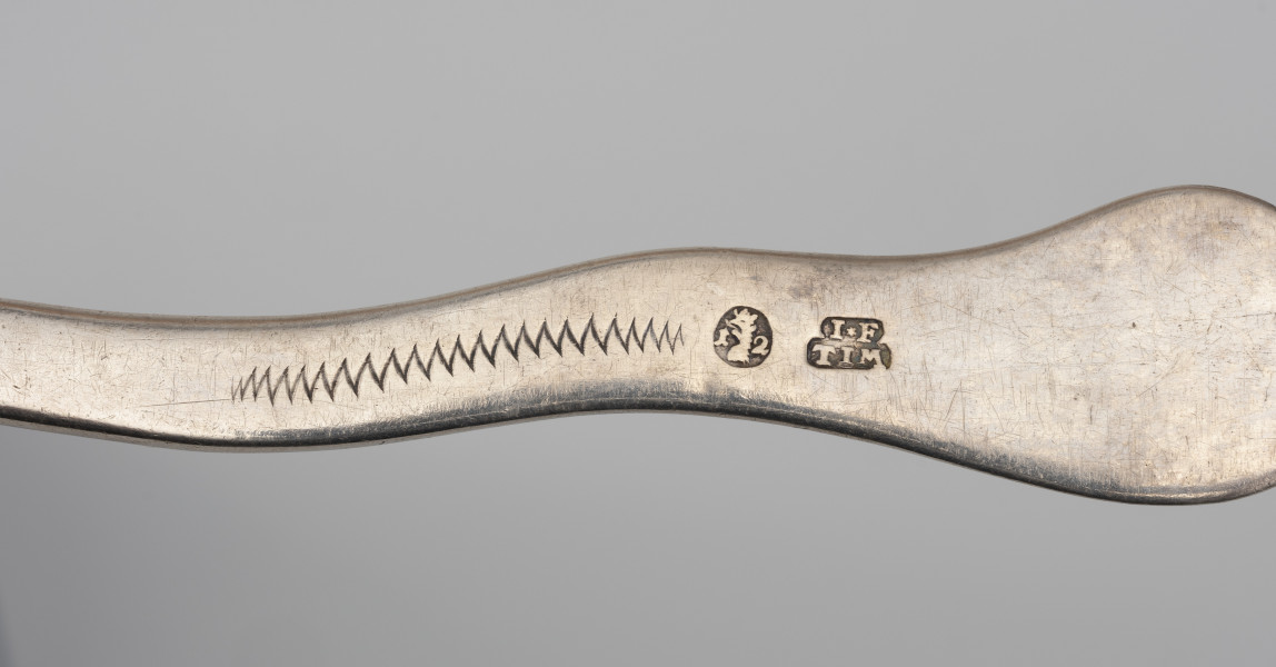 łyżka wazowa - detal; fragment trzonka łyżki wazowej; od prawej napis I*F | TIM - cecha warsztatowa mistrza, obok w owalnym polu głowa gryfa w koronie pomiędzy cyframi, z lewej grawerowany szlaczek w kształcie zygzaka