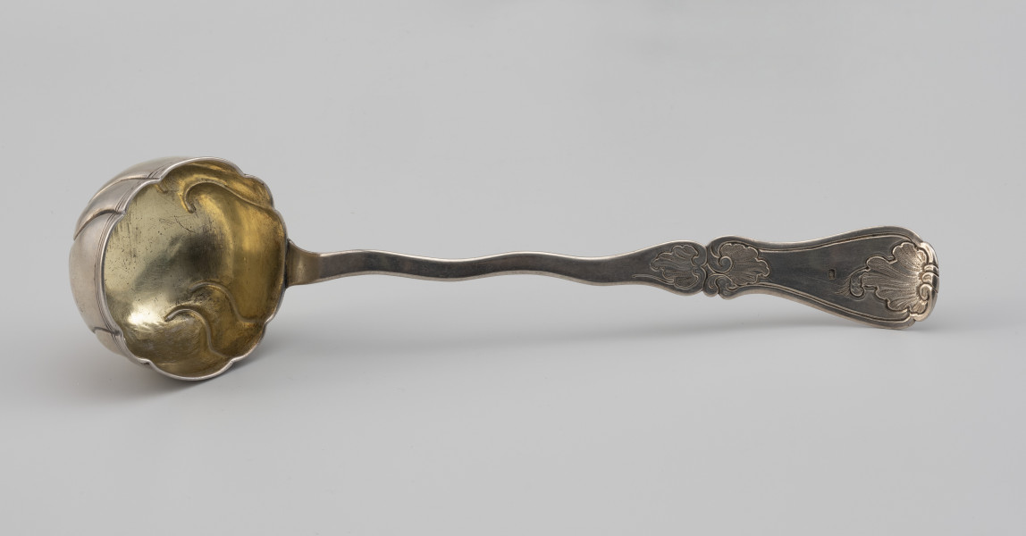 łyżka wazowa - Ujęcie z góry; Rokokowa łyżka wazowa o puklowanej powierzchni i falistym brzegu, z rytowaną podwójną linią przy krawędzi. Pukle wydzielone czterema parami repusowanych esowatych linii. Wnętrze czerpaka złocone. Trzonek płaski, o asymetrycznym, falistym wykroju. Uchwyt wydzielony przewężeniem, na końcu rozszerzający się i wygięty lekko w górę, zdobiony grawerowaną i fakturowaną rokokową dekoracją. Na uchwycie znak FW w prostokatnym polu - pruska cecha kontrybucyjna z l. 1809–1812 dla sreber wykupionych przez właściciela
