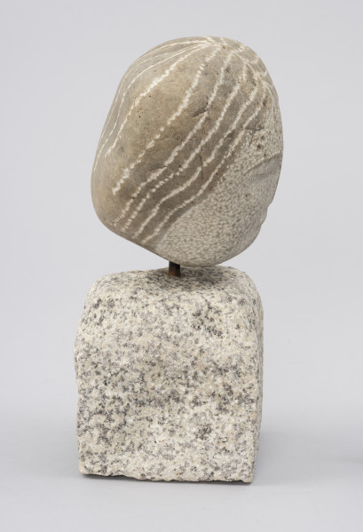 rzeźba; Prasłowianka I - ujęcie z lewej; Owalna bryła naturalnego kamienia polnego osadzona na kostce kamiennej. Skała tworząca głowę przekształcona w niewielkim stopniu. Wydobyto cechy fryzury - punktowym rytowaniem zaznaczony został przedziałek po środku głowy oraz linie wyznaczające szerokie pasma prostych, ciemnych włosów, biegnące od przedziałka do tyłu głowy, sugerując związanie w koński ogon.