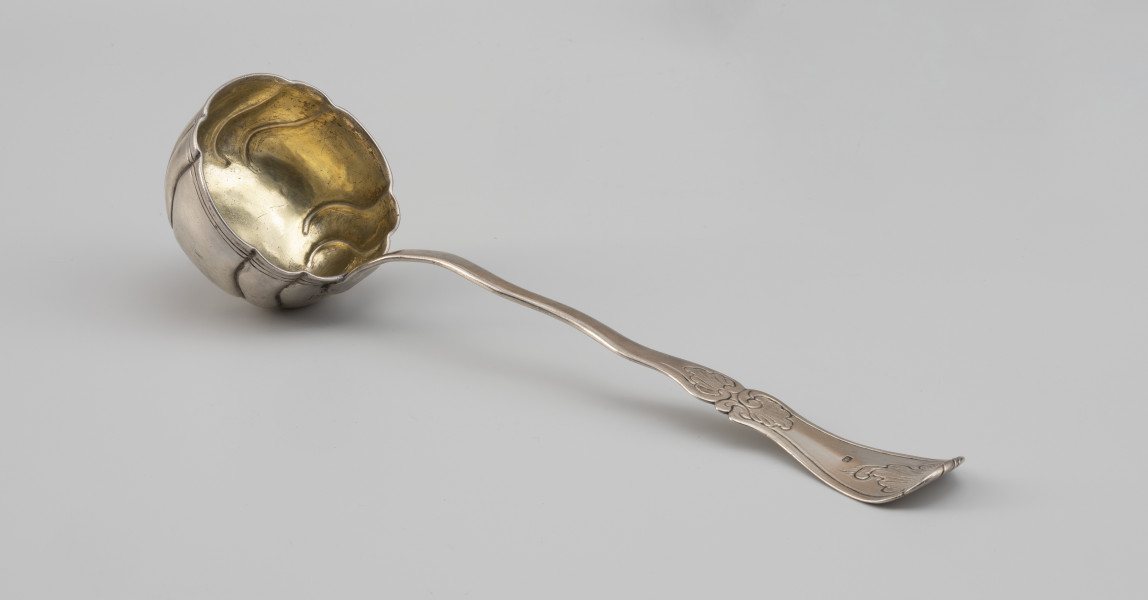 łyżka wazowa - Ujęcie ze skosu; Rokokowa łyżka wazowa o puklowanej powierzchni i falistym brzegu, z rytowaną podwójną linią przy krawędzi. Pukle wydzielone czterema parami repusowanych esowatych linii. Wnętrze czerpaka złocone. Trzonek płaski, o asymetrycznym, falistym wykroju. Uchwyt wydzielony przewężeniem, na końcu rozszerzający się i wygięty lekko w górę, zdobiony grawerowaną i fakturowaną rokokową dekoracją.