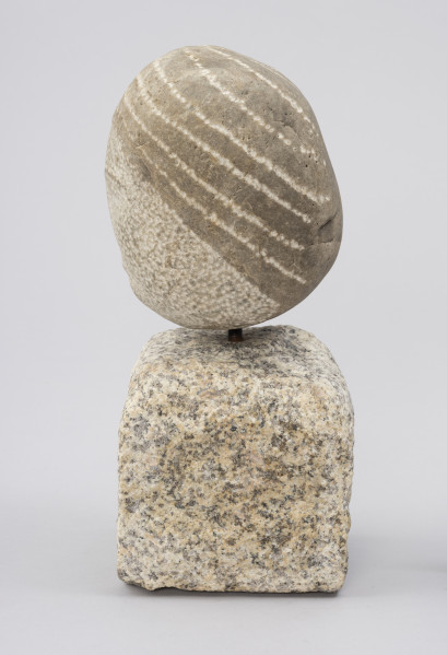 rzeźba; Prasłowianka I - ujęcie z prawej; Owalna bryła naturalnego kamienia polnego osadzona na kostce kamiennej. Skała tworząca głowę przekształcona w niewielkim stopniu. Wydobyto cechy fryzury - punktowym rytowaniem, linie wyznaczające szerokie pasma prostych, ciemnych włosów, biegnące od przedziałka do tyłu głowy, sugerując związanie w koński ogon.