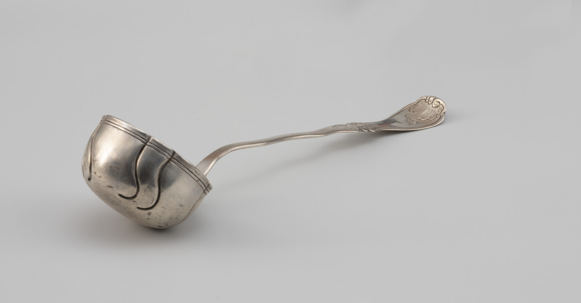 łyżka wazowa - Ujęcie z boku; Rokokowa łyżka wazowa o puklowanej powierzchni i falistym brzegu, z rytowaną podwójną linią przy krawędzi. Pukle wydzielone czterema parami repusowanych esowatych linii. Trzonek płaski, o asymetrycznym, falistym wykroju. Uchwyt wydzielony przewężeniem, na końcu rozszerzający się i wygięty lekko w górę, zdobiony grawerowaną i fakturowaną rokokową dekoracją.