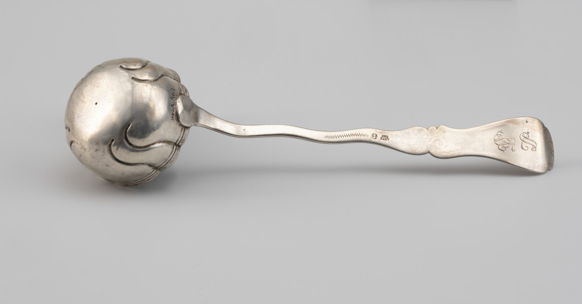 łyżka wazowa - Ujęcie z dołu; Rokokowa łyżka wazowa o puklowanej powierzchni i falistym brzegu, z rytowaną podwójną linią przy krawędzi. Pukle wydzielone czterema parami repusowanych esowatych linii. Trzonek płaski, o asymetrycznym, falistym wykroju. Uchwyt wydzielony przewężeniem, na końcu rozszerzający się i wygięty lekko w górę. Na środku w owalnym polu głowa gryfa w koronie pomiędzy cyframi, obok z prawej cecha warsztatowa mistrza I*F TIM