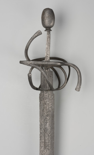 rapier - Ujęcie 3/4; Rapier o głowni prostej, obusiecznej, z ricassem. Na ricassie i krótkim odcinku głowni biegnie zaznaczone dwoma korytkami zbrocze. Rękojeść ma krzyżowy jelec w esowatej formie i rozbudowany kosz do ochrony dłoni. Kosz składa się z trzech obłęków bocznych rozchodzących się ku ostrzu i osłaniających ricasso, po drugiej stronie są dwa obłęki boczne, z których górny jest większy, połączonych „oślą podkową” i dodatkowo dwoma obłękami. Głowica jest owalna, wydłużona z wyodrębnioną podstawą w formie pierścieni, ozdobiona jest wzdłużnie nacięciami i zakuta od góry.