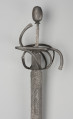 rapier - Ujęcie 3/4; Rapier o głowni prostej, obusiecznej, z ricassem. Na ricassie i krótkim odcinku głowni biegnie zaznaczone dwoma korytkami zbrocze. Rękojeść ma krzyżowy jelec w esowatej formie i rozbudowany kosz do ochrony dłoni. Kosz składa się z trzech obłęków bocznych rozchodzących się ku ostrzu i osłaniających ricasso, po drugiej stronie są dwa obłęki boczne, z których górny jest większy, połączonych „oślą podkową” i dodatkowo dwoma obłękami. Głowica jest owalna, wydłużona z wyodrębnioną podstawą w formie pierścieni, ozdobiona jest wzdłużnie nacięciami i zakuta od góry.