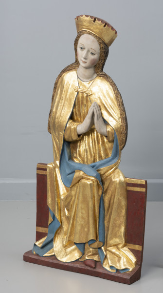 dwufigurowa grupa płaskorzeźb ołtarzowych - Ujęcie przodu ze skosu z prawej; Płaskorzeźbiona figura przedstawia na szerokiej ławie tronuje Maria. Postać ubrana jest w długą tunikę przewiązaną w pasie i płaszcz spięty klamrą powyżej piersi. Miękka, lejąca się ku podstawie rzeźby złocista tkanina, szata połyskuje efektownie marszczeniami o łagodnych grzbietach. Na głowie wysoka złocona korona o gładkiej obręczy, zwieńczona zygzakiem. Maria lekko przechyla głowę wlewo, dłonie trzymając w geście modlitewnym przy piersi. Jej młodzieńczą twarz o wysokim czole i łagodnych rysach otaczają pasma długich falujących, złocistych włosów.  Długie włosy opadają na ramiona. Tył rzeźby opracowany płasko. Fleurony wieńczące obręcze koron nie zachowały się. Złocenia płaskorzeźby częściowo rekonstruowane.