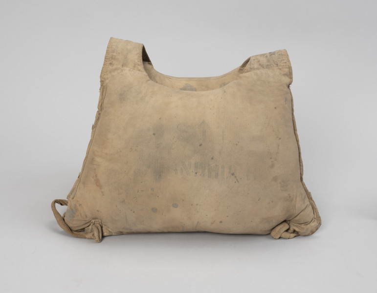 kamizelka ratunkowa - Ujęcie z przodu; Kamizelka wykonana z dwóch pływaków płóciennych, przednim i tylnym, wypełnionych trawą morską, o kształtach zbliżonych do prostokąta lub trapezu (64 x 40 cm). Górne narożniki pływaków połączone ze sobą pustymi komorami umożliwiającymi swobodne zwisanie dwóch pozostałych komór-pływaków, tworząc jednocześnie otwór na środku na głowę o średnicy 18 cm. Przy dolnych narożnikach kamizelki przyszyte przelotki z taśmy parcianej. W obydwu parach przelotek, wzdłuż dłuższych boków umieszczono taśmy parciane o długości 9 cm, zakończone stoperami powstałymi ze złożenia i przeszycia końcówek taśm, które zapobiegają wysunięciu się taśm z przelotek. Na jednym z dłuższych boków, przy szwach naszyte dodatkowe kawałki tkaniny wzmacniające naderwane szwy. Słabo czytelny czarny nadruk.