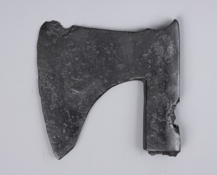 żeleźce topora, oksza typu X (Głosek) - Ujęcie z przodu; Oksza w typie X (według typologii Mariana Głoska), o górnej krawędzi wznoszącej się lekko ku górze, dolnej opadającej łukiem. Broda lekko ścięta pod skosem, szyja szeroka. Ostrze jest asymetryczne i lekko wygięte. Osada jest długa, zakuta od góry, z płynnie wyprofilowanym zgrubieniem w górnej krawędzi przechodzącym w wystający kolec. Osada ma ubytki metalu spowodowane korozją. Wewnątrz osady zachowały się drewniane pozostałości trzonka.