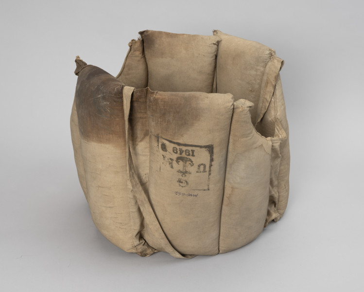 kamizelka ratunkowa trawiasta - Ujęcie z przodu; Kamizelka wykonana z dwóch kawałków płótna beżowego koloru o kształtach zbliżonych do prostokąta ( 135x x 38 cm) złożona z ośmiu komór wypornościowych, cztery o wymiarach 17,5 x 38, cztery o wymiarach 10x38 z wyprofilowanym wycięciem na głowę, dodatkowo dwie komory puste o wymiarach 26x 8 cm umożliwiające swobodne zwisanie sąsiednich komór, na wysokości 19 centymetrów wszyte wzmocnienie z pasów parcianych, w połowie wysokości pływaków skrajnych przyszyte taśmy parciane o wymiarach długość 110 cm x szerokość 2cm, na piątej komorze znak stempla o wym. 10 x 10 cm w kolorze czarnym w miejscu centralnym symbol kotwicy obwiedziony literami UMG , poniżej pięty kotwicy cyfry 1949, pływaki wypełnione trawą morską. Krawędzie płócien zszyte ze sobą. Do dwóch mniejszych pływaków z wycięciem na głowę przyszyta parciana przelotka o wym. 46 x 2 cm. Jedna z pustych komór uszkodzona- naderwany materiał nad wszytym wzmocnieniem kieszeni. Całość nosi ślady eksploatacji z widocznymi tłustymi plamami.