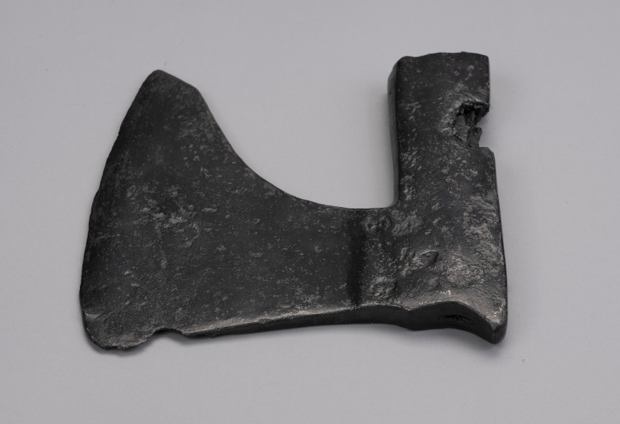 żeleźce topora, oksza typu X (Głosek) - Ujęcie lekko z góry; Oksza w typie X (według typologii Mariana Głoska), o górnej krawędzi wznoszącej się lekko ku górze, dolnej opadającej łukiem. Broda lekko ścięta pod skosem, szyja szeroka. Ostrze jest asymetryczne i lekko wygięte. Osada jest długa, zakuta od góry, z płynnie wyprofilowanym zgrubieniem w górnej krawędzi przechodzącym w wystający kolec. Osada ma ubytki metalu spowodowane korozją. Wewnątrz osady zachowały się drewniane pozostałości trzonka.