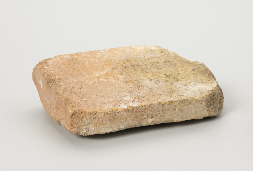 cegła szamotowa - Ujęcie przodu ze skosu z lewej; Prostopadłościenna cegła z jasnej glinki szamotowej. Prawa strona obtłuczona. Cegła niejednolita, ziarnista. W centrum płaszczyzny ścianki poziomej tłoczony, wykonany metodą stempla, wklęsły napis DIDIER 100.