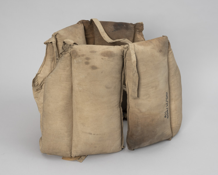 kamizelka ratunkowa trawiasta - Ujęcie z przodu; Kamizelka wykonana z dwóch kawałków płótna beżowego koloru o kształtach zbliżonych do prostokąta ( 135x x 38 cm) złożona z ośmiu komór wypornościowych, cztery o wymiarach 17,5 x 38, cztery o wymiarach 10x38 z wyprofilowanym wycięciem na głowę, dodatkowo dwie komory puste o wymiarach 26x 8 cm umożliwiające swobodne zwisanie sąsiednich komór, na wysokości 19 centymetrów wszyte wzmocnienie z pasów parcianych, w połowie wysokości pływaków skrajnych przyszyte taśmy parciane o wymiarach długość 110 cm x szerokość 2cm, pływaki wypełnione trawą morską. Krawędzie płócien zszyte ze sobą. Całość nosi ślady eksploatacji z widocznymi tłustymi plamami.