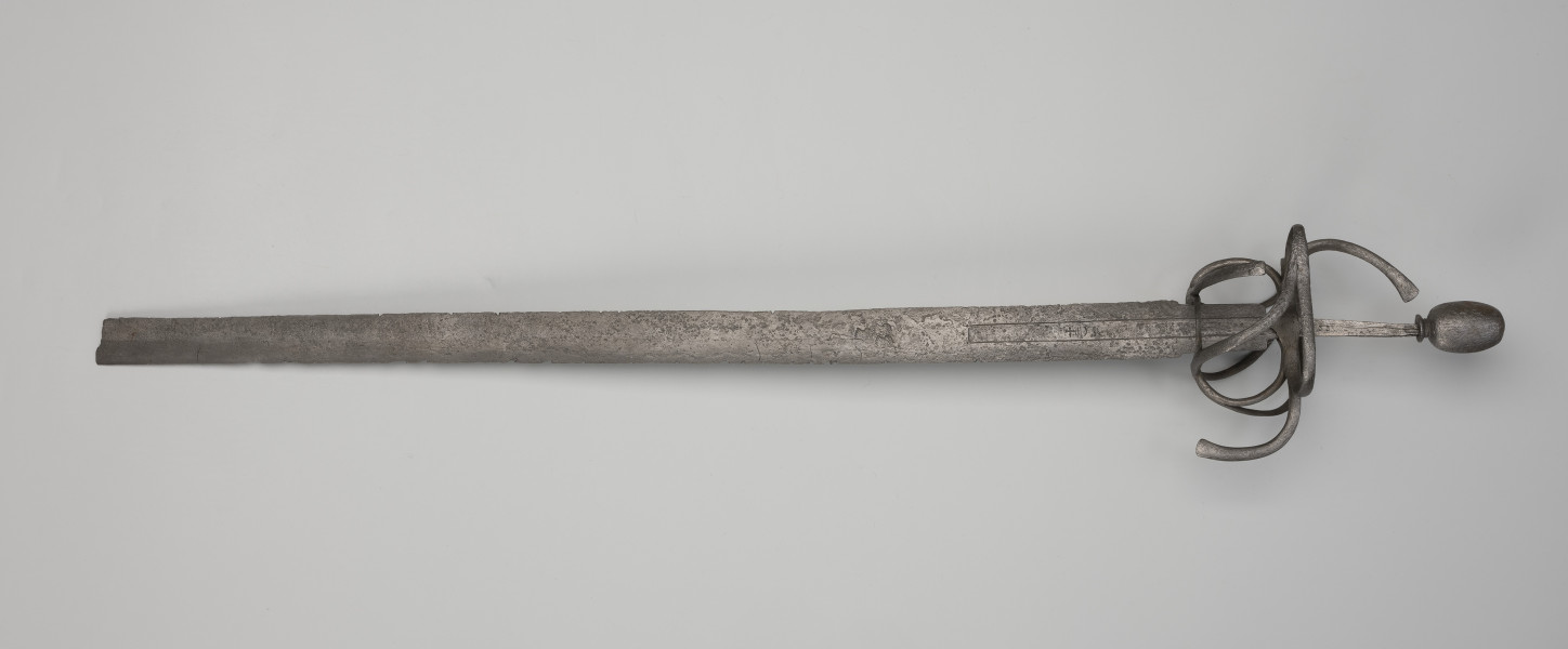 rapier - Ujęcie z tyłu; Rapier o głowni prostej, obusiecznej, z ricassem. Na ricassie i krótkim odcinku głowni biegnie zaznaczone dwoma korytkami zbrocze. Sztych jest ułamany. Rękojeść ma krzyżowy jelec w esowatej formie i rozbudowany kosz do ochrony dłoni. Kosz składa się z trzech obłęków bocznych rozchodzących się ku ostrzu i osłaniających ricasso, po drugiej stronie są dwa obłęki boczne, z których górny jest większy, połączonych „oślą podkową” i dodatkowo dwoma obłękami. Trzon zwęża się ku końcowi, jego okładzina nie zachowała się. Głowica jest owalna, wydłużona z wyodrębnioną podstawą w formie pierścieni, ozdobiona jest wzdłużnie nacięciami i zakuta od góry.