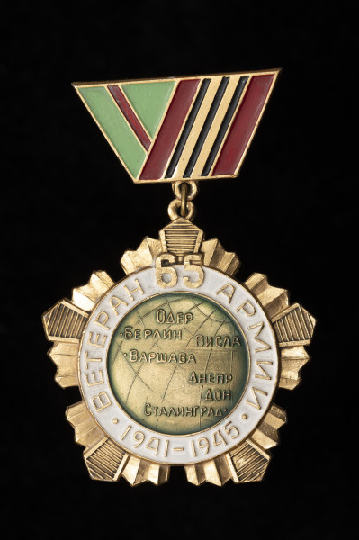 Weteran 65 Armii - Ujęcie z przodu; Odznaka jednostronnego medalu w formie krążka na pięciobocznej plakiecie z zawieszką w formie złożonej wstążki orderowej. W centrum okrągłej odznaki wyobrażona została kula ziemska z detalami kartograficznymi oraz nazwami miejscowości i rzek w języku rosyjskim. Na otoku pokrytym białą emalią wytłoczony napis rosyjski cyrylicą: WETERAN 65 ARMII 1941-1945. Od spodu odznakę uzupełnia pięcioboczna plakieta przybierająca postać rozchodzących się promieni. Promienie ułożone są w pięć grup po pięć, gdzie środkowy promień każdej grupy stanowi narożnik pięcioboku i zaznaczony jest prążkowaną fakturą. Trapezowata, emaliowana zawieszka ma formę ukośnie złożonych wstążek orderowych: zielono-czerwonej po lewej i czerwono-złoto-czarnej po prawej stronie medalu. Zawieszka wyposażona jest w zaczep agrafkowy, który umożliwia przymocowanie medalu do ubrania lub innej tkaniny. Szczyt plakiety oraz dół zawieszki zaopatrzone są w metalowe kółka, które spaja pojedyncze, okrągłe ogniwo. Części nie pokryte kolorowymi emaliami zostały pomalowane na złoty odcień.