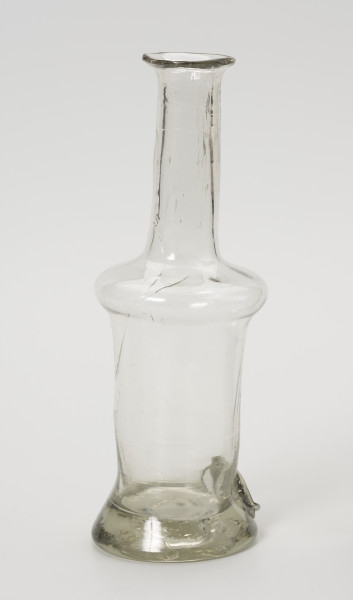 karafinka - Ujęcie z tyłu ze skosu z lewej; Niewielka butelka o pojemności 0,25 L. Wydmuchana na piszczelu z bezbarwnego szkła – na brzuścu widoczne smugi z nie do końca prawidłowego procesu produkcji. Kształt podłużny, wąski, o grubym, wyraźnie wyodrębnionym dnie, brzusiec w górnej części uwypuklony przechodzi w wąską, długą szyjkę zakończoną płasko wywiniętym niewielkim wylewem. Korka brak.