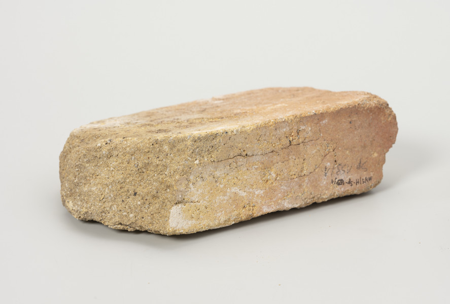 cegła szamotowa - Ujęcie tyłu; Prostopadłościenna cegła z jasnej glinki szamotowej. Lewa strona obtłuczona. Cegła niejednolita, ziarnista.