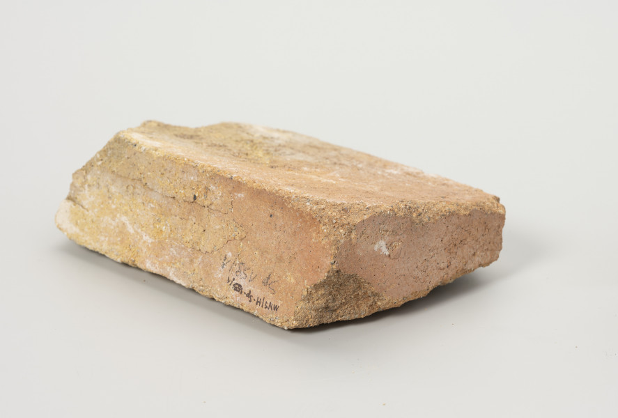 cegła szamotowa - Ujęcie z tyłu; Prostopadłościenna cegła z jasnej glinki szamotowej. Lewa strona obtłuczona. Cegła niejednolita, ziarnista.