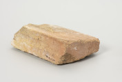 cegła szamotowa - Ujęcie z tyłu; Prostopadłościenna cegła z jasnej glinki szamotowej. Lewa strona obtłuczona. Cegła niejednolita, ziarnista.