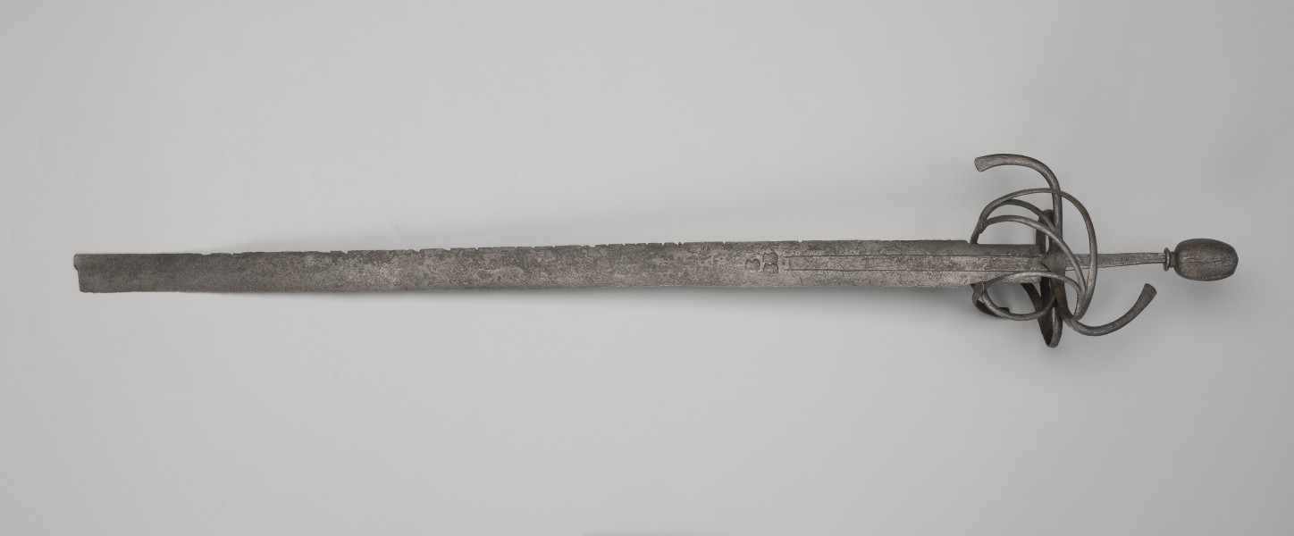 rapier - Ujęcie z przodu; Rapier o głowni prostej, obusiecznej, z ricassem. Na ricassie i krótkim odcinku głowni biegnie zaznaczone dwoma korytkami zbrocze. Sztych jest ułamany. Rękojeść ma krzyżowy jelec w esowatej formie i rozbudowany kosz do ochrony dłoni. Kosz składa się z trzech obłęków bocznych rozchodzących się ku ostrzu i osłaniających ricasso, po drugiej stronie są dwa obłęki boczne, z których górny jest większy, połączonych „oślą podkową” i dodatkowo dwoma obłękami. Trzon zwęża się ku końcowi, jego okładzina nie zachowała się. Głowica jest owalna, wydłużona z wyodrębnioną podstawą w formie pierścieni, ozdobiona jest wzdłużnie nacięciami i zakuta od góry.