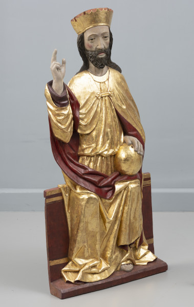 dwufigurowa grupa płaskorzeźb ołtarzowych - Ujęcie przodu ze skosu z lewej; Płaskorzeźbiona figura, na szerokiej ławie tronuje Chrystus. Postać ubrana jest w długą tunikę przewiązaną w pasie, płaszcz spięty klamrą powyżej piersi. Miękka, lejąca się ku podstawie rzeźby złocista tkanina,  szata połyskuje efektownie marszczeniami o łagodnych grzbietach. Na głowie wysoka złocona korona o gładkiej obręczy, zwieńczona zygzakiem. Chrystus swoją prawą rękę podnosi w geście błogosławieństwa, lewą opiera na złotej kuli trzymanej na kolanie. Jego pociągłą twarz o wyraźnie zaznaczonych kościach policzkowych pokrywa ciemny zarost. Długie włosy opadają na ramiona. Widoczne pęknięcie drewna na twarzy Chrystusa wzdłuż jego prawej skroni, poprzez policzek w kierunku ust. Fleurony wieńczące obręcze koron nie zachowały się. Złocenia płaskorzeźby częściowo rekonstruowane.