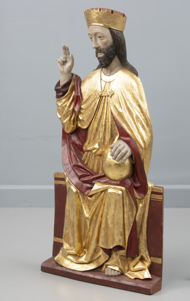 dwufigurowa grupa płaskorzeźb ołtarzowych - Ujęcie przodu ze skosu z prawej; Płaskorzeźbiona figura, na szerokiej ławie tronuje Chrystus. Postać ubrana jest w długą tunikę przewiązaną w pasie, płaszcz spięty klamrą powyżej piersi. Miękka, lejąca się ku podstawie rzeźby złocista tkanina,  szata połyskuje efektownie marszczeniami o łagodnych grzbietach. Na głowie wysoka złocona korona o gładkiej obręczy, zwieńczona zygzakiem. Chrystus swoją prawą rękę podnosi w geście błogosławieństwa, lewą opiera na złotej kuli trzymanej na kolanie. Jego pociągłą twarz o wyraźnie zaznaczonych kościach policzkowych pokrywa ciemny zarost. Długie włosy opadają na ramiona. Widoczne pęknięcie drewna na twarzy Chrystusa wzdłuż jego prawej skroni, poprzez policzek w kierunku ust. Fleurony wieńczące obręcze koron nie zachowały się. Złocenia płaskorzeźby częściowo rekonstruowane.