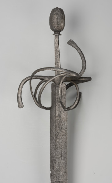 rapier - Ujęcie 3/4; Rapier o głowni prostej, obusiecznej, z ricassem. Na ricassie i krótkim odcinku głowni biegnie zaznaczone dwoma korytkami zbrocze. Rękojeść ma krzyżowy jelec w esowatej formie i rozbudowany kosz do ochrony dłoni. Kosz składa się z trzech obłęków bocznych rozchodzących się ku ostrzu i osłaniających ricasso, po drugiej stronie są dwa obłęki boczne, z których górny jest większy, połączonych „oślą podkową” i dodatkowo dwoma obłękami. Głowica jest owalna, wydłużona z wyodrębnioną podstawą w formie pierścieni, ozdobiona jest wzdłużnie nacięciami i zakuta od góry.