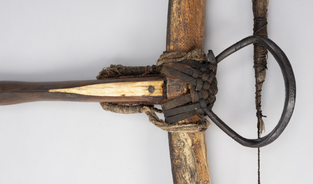 kusza - Ujęcie fragmentu; fragment środka kuszy średniowiecznej.  Od góry na łożu zamocowana jest kościana płytka z wgłębieniem na prowadnicę bełta przytwierdzona w najszerszej części gwoździem. W przedniej części łoża po boku widoczne są otwory z pozostałością splotu mocującego łuczywo dalej ku końcowi widoczne są nity mocujące spust. Łuczysko jest mocowane do frontu łoża wspomnianym oplotem. Jest pokryte korą drzewa brzozowego. Pośrodku łuku przywiązane jest rzemieniem żelazne strzemię, zdobione po bokach nacięciami.