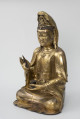 Bodhisattwa Guanyin - ujęcie przodu ze skosu z prawej; Duża, wykonana ze złoconego brązu figura przedstawia siedzącą w pozie medytacyjnej kobietę Bodhisattwę Guanyin (chin. 观音, sanskr. Awalokiteśwara, tyb. Czenrezig). Postać ma spuszczone, przymknięte oczy, symetryczną, pełną twarz o pogodnym i spokojnym wyrazie oraz gładko zaczesane włosy, schowane pod diademem i zaciągniętą na głowę tkaniną. Ubrana jest w długie, warstwowe, jedwabne szaty królewskie oraz wyszukaną, ciężką biżuterię książęcą: diadem, w którego centrum znajduje się niebiański Budda Amitabha (sanskr. Amitābha); pary dużych, okrągłych kolczyków ze spiczastymi zawieszkami oraz grubego naszyjnika z klejnotem, od którego odchodzi siedem pasów korali z wisiorkami. Jej prawa dłoń uniesiona na wysokości klatki piersiowej ułożona jest pionowo, jej środkowy palec i kciuk tworzą okrąg trzymając niezidentyfikowany, haczykowaty przedmiot. Poniżej znajduje się ustawiona poziomo lewa dłoń, która ściska niewielką wazę.