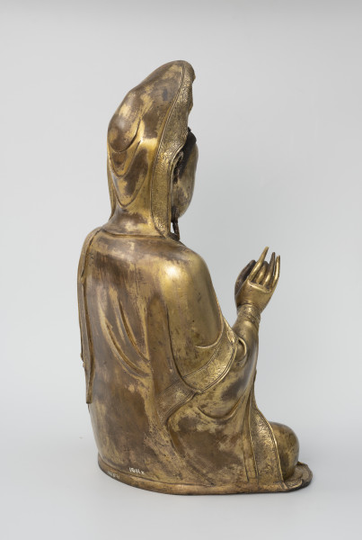 Bodhisattwa Guanyin - ujęcie tyłu ze skosu z prawej; Duża, wykonana ze złoconego brązu figura przedstawia siedzącą w pozie medytacyjnej kobietę Bodhisattwę Guanyin (chin. 观音, sanskr. Awalokiteśwara, tyb. Czenrezig). Postać ma spuszczone, przymknięte oczy, symetryczną, pełną twarz o pogodnym i spokojnym wyrazie oraz gładko zaczesane włosy, schowane pod diademem i zaciągniętą na głowę tkaniną. Ubrana jest w długie, warstwowe, jedwabne szaty królewskie. Jej prawa dłoń uniesiona na wysokości klatki piersiowej ułożona jest pionowo, jej środkowy palec i kciuk tworzą okrąg. Z tyłu figury, przy dolnej krawędzi biały napis pod postacią numeru: 1011z.