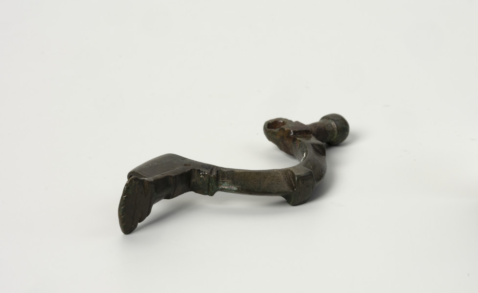 fibula z brązu z Borkowic typu Bügelknopffibel - Ujęcie z góry; Zapinka z brązu z guzkiem na łukowatym kabłąku typu Bügelknopffibel. Fibula pozbawiona jest sprężynki i szpili. Kabłąk jest zdobiony profilowanymi guzkami w symetrycznym układzie.