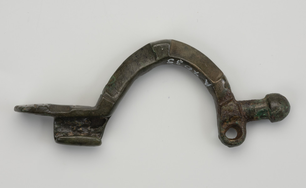 fibula z brązu z Borkowic typu Bügelknopffibel - Ujęcie z boku; Zapinka z brązu z guzkiem na łukowatym kabłąku typu Bügelknopffibel. Fibula pozbawiona jest sprężynki i szpili.