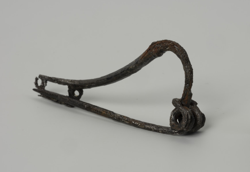 fibula wariant N wg Kostrzewskiego - Ujęcie przodu ze skosu z lewej; Żelazna zapinka wariantu N wg Kostrzewskiego, z wysokim kabłąkiem z okrągłego drutu, z guzem plastycznym. Sprężynka z czterema zwojami i dolną cięciwą. Ażurowa pochewka ma dwa otwory.