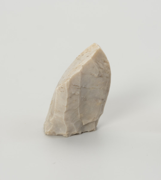 rdzeń stożkowy - Ujęcie z boku; Rdzeń z bryły krzemienia kredowego bardzo wysokiej jakości w końcowym stadium eksploatacji o znacznym stopniu redukcji uniemożliwiającym jego utrzymanie. Forma zbliżona do stożka lub ostrosłupa o dookolnej odłupni i płaskiej, lekko wklęsłej pięcie uformowanej jednym odbiciem. Widoczny na jednej ze stron duży negatyw wskazuje na naprawę rdzenia krótko przed zakończeniem eksploatacji. W masie widoczne są liczne długie egzoszkielety organizmów morskich, ułożone w podłużne linie. Zabytek w całości pokryty jest białą patyną wskazującą na długotrwałe zaleganie na powierzchni.